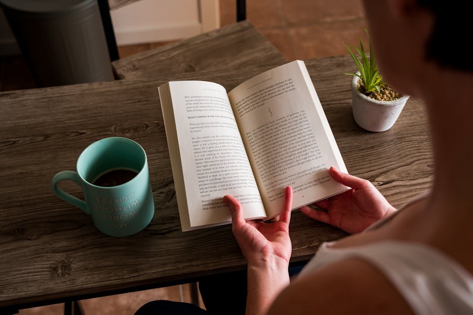 person reading book at cafe table with coffee and laptop
